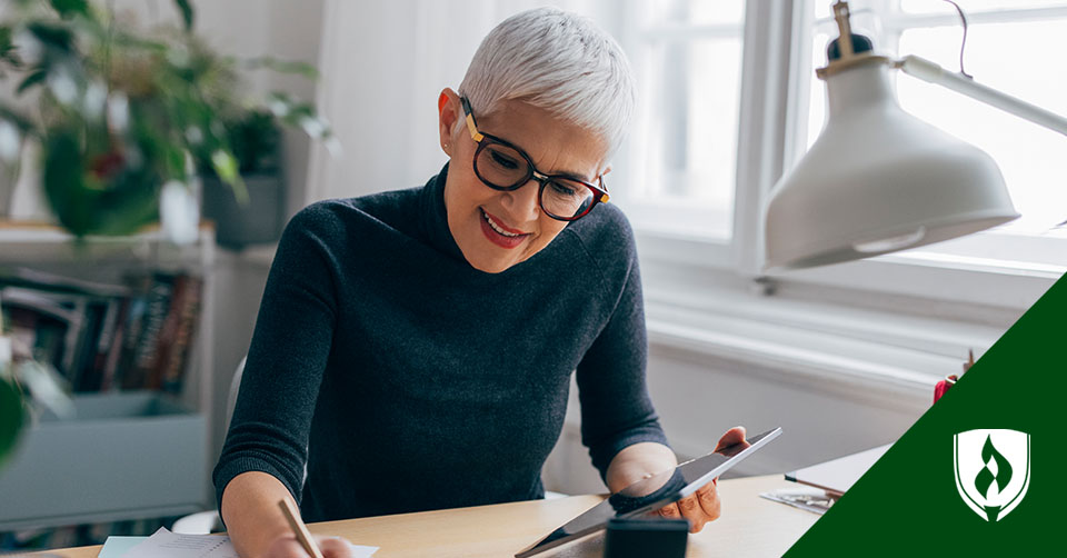 A white haired woman in a black sweater sits at her desk looking at her tablet and writing something