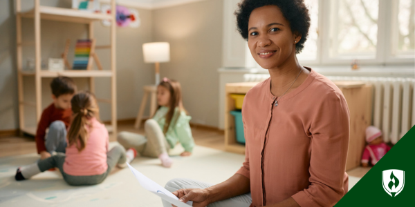 An early childhood educator looks at the camera with three young kids playing in the background