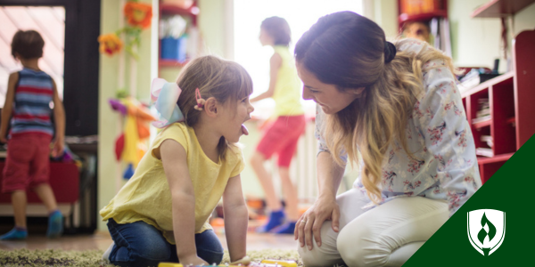 A day care provider plays with a little girl in a yellow shirt as other kids play in the background of her living room