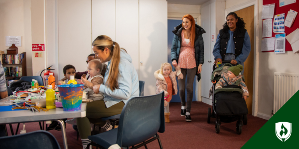 Two moms walk into a childcare center where a teacher plays with a toddler