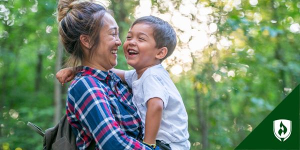 An educator holds a child, laughing with him in the forest