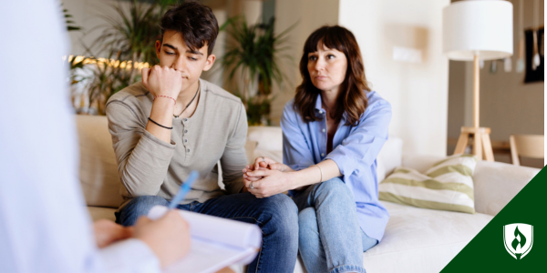 A teenager holds his mom's hand in an office, looking stricken