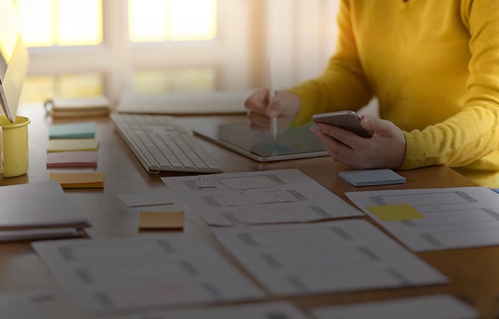 person writing on a tablet with mobile device in other hand on table with mockups and sticky notes