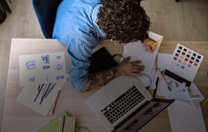 overhead view of person writing on desk next to laptop with art supplies nearby